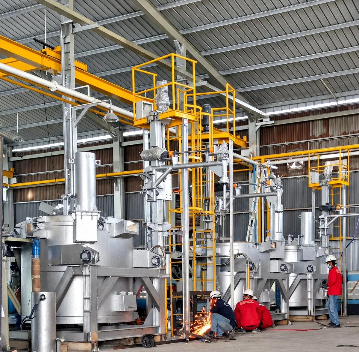 MWT crucible melting and holding furnaces on the workshop floor with yellow access platforms, gas piping and technicians at work